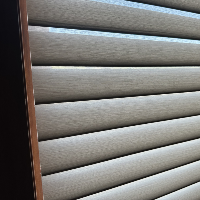 A close-up view of horizontal window blinds with a textured taupe surface, set within a dark wooden frame. The image highlights the interplay of light and shadow across the slats, with a bright background visible through small gaps.